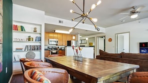 A spacious dining area with a wooden table and leather chairs, a kitchen in the background, and a bookshelf with books and decorative items.