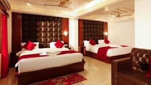 A hotel room with two beds, each with a dark headboard, red pillows, and white bedding. There is a wooden floor, a red carpet, and a brown leather chair. - AT Hotels & Resorts (Port Blair)