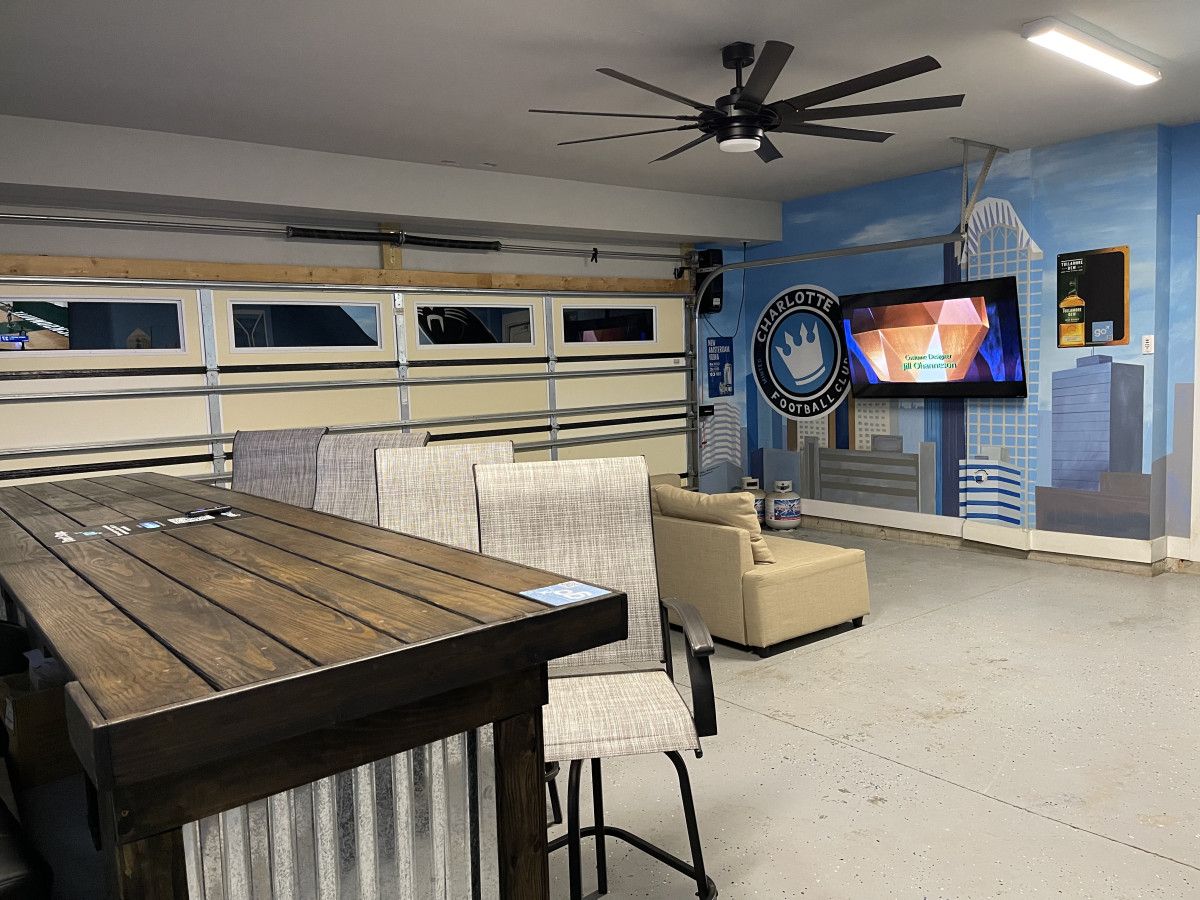 Une pièce avec une table en bois, des chaises, un ventilateur de plafond, un téléviseur et un mur orné d’une fresque représentant un paysage urbain.