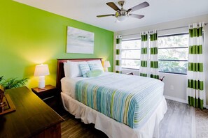 Un dormitorio con cama, mesita de noche, lámpara, ventilador de techo y ventana con cortinas.