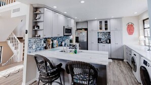 Una cocina moderna con armarios grises, una isla central, una lavadora y un comedor con sillas de mimbre.