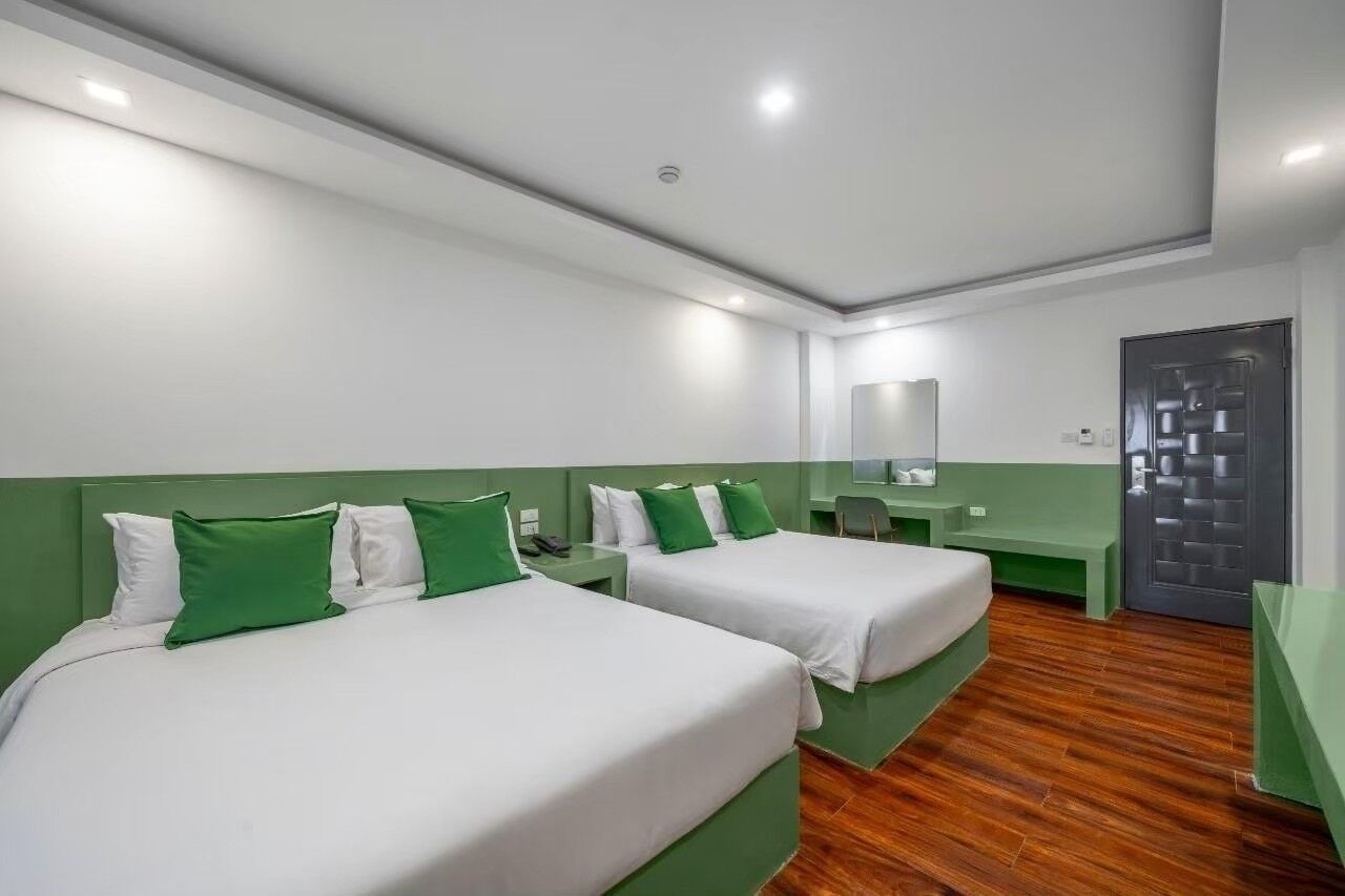 A hotel room with two beds, green headboards, white bedding, a wooden floor, a mirror, and a door with a geometric pattern.