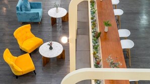 Un área de descanso moderna con una mesa de madera, sillas amarillas, un sofá azul y pequeñas mesas redondas con objetos decorativos.