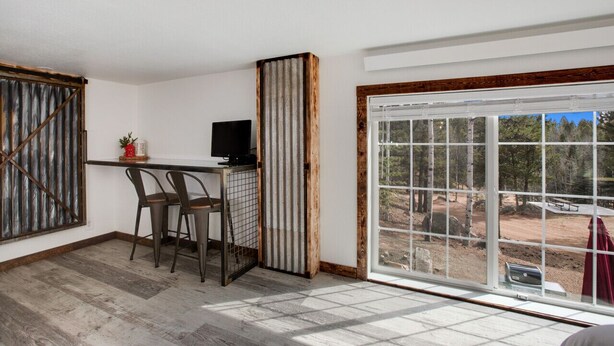 Ein Zimmer mit Schreibtisch, zwei Stühlen, einem Fernseher und einem großen Fenster mit Blick auf eine bewaldete Landschaft.
