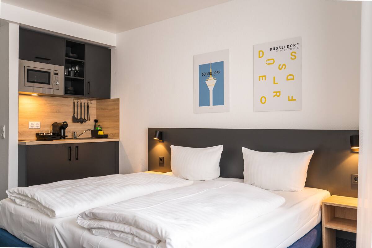 A modern hotel room with a bed, a built-in kitchenette, and a wall-mounted artwork.
