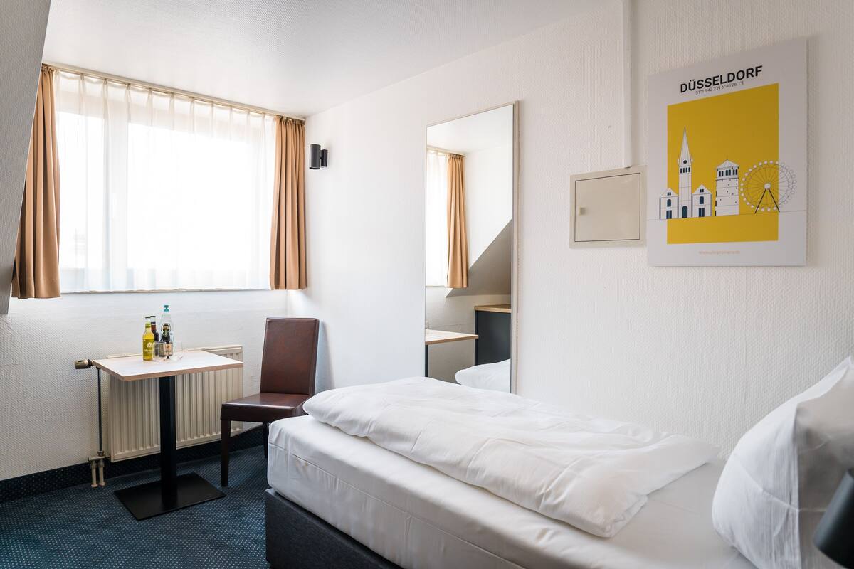 A hotel room with a bed, a desk, a chair, a window with curtains, a mirror, and a poster on the wall.
