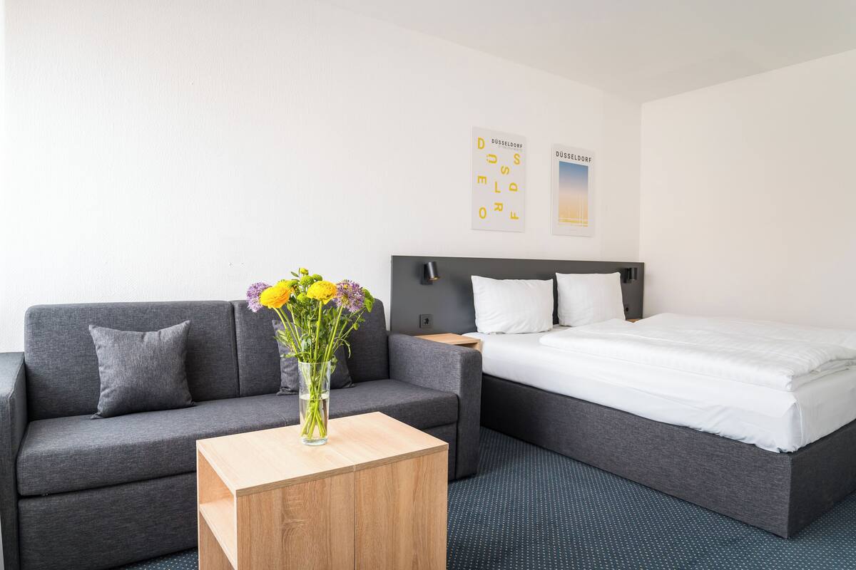 A hotel room with a grey sofa, a wooden side table, a bed with white bedding, and a vase with flowers.