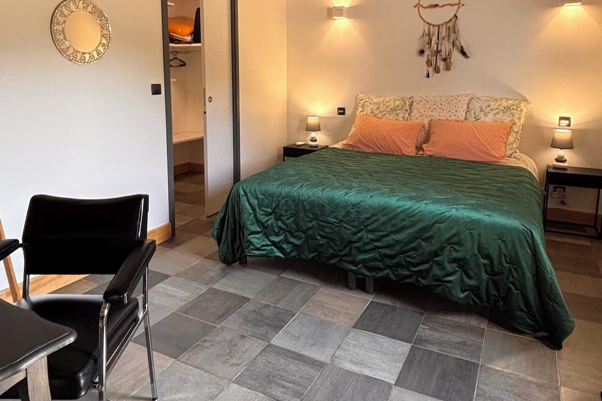 A bedroom with a green bedspread, a black chair, a small table, and a wardrobe.