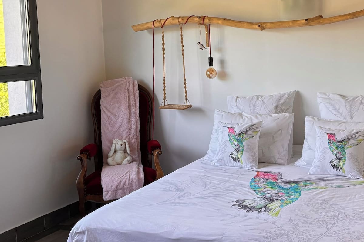 A bedroom with a bed, a bench, a chair, a hanging light fixture, and a window.