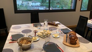 Een eettafel gedekt met diverse kazen, brood en een stokbrood op een houten plank, met een mooi uitzicht door een groot raam.