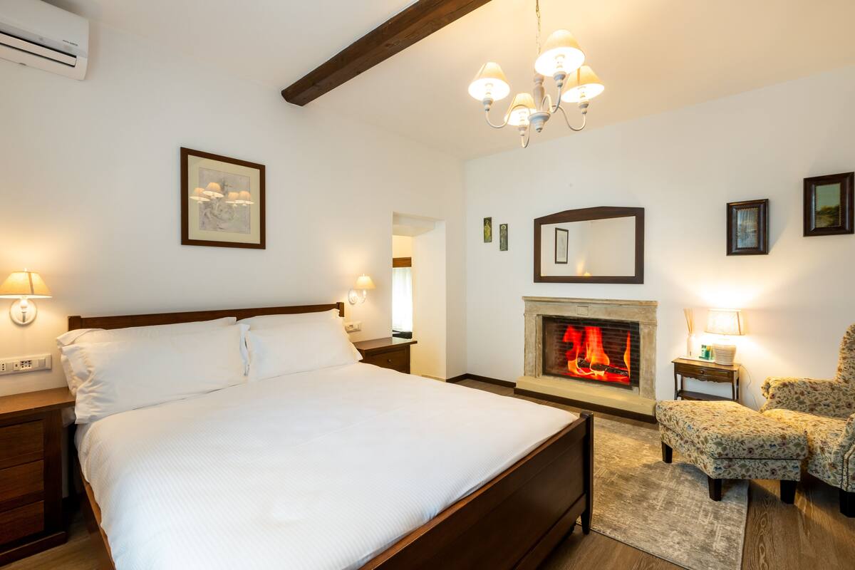 A bedroom with a large bed, a fireplace, a chandelier, and a chair.