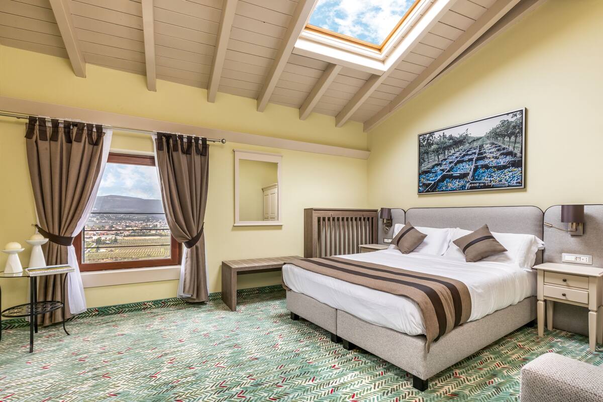 A bedroom with a large bed, a bedside table, a nightstand, a window with curtains, a skylight, and a painting on the wall.