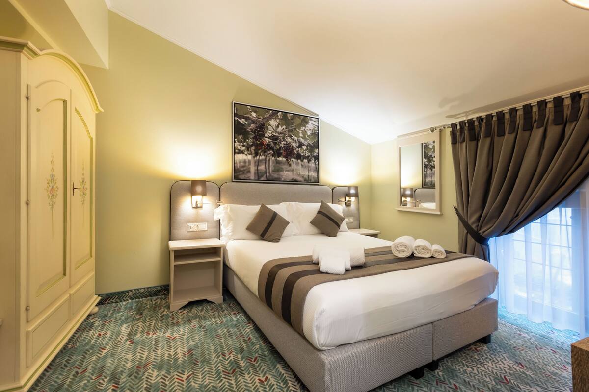 A hotel room with a large bed, two bedside tables, a headboard, a painting, a window with curtains, and a patterned carpet.