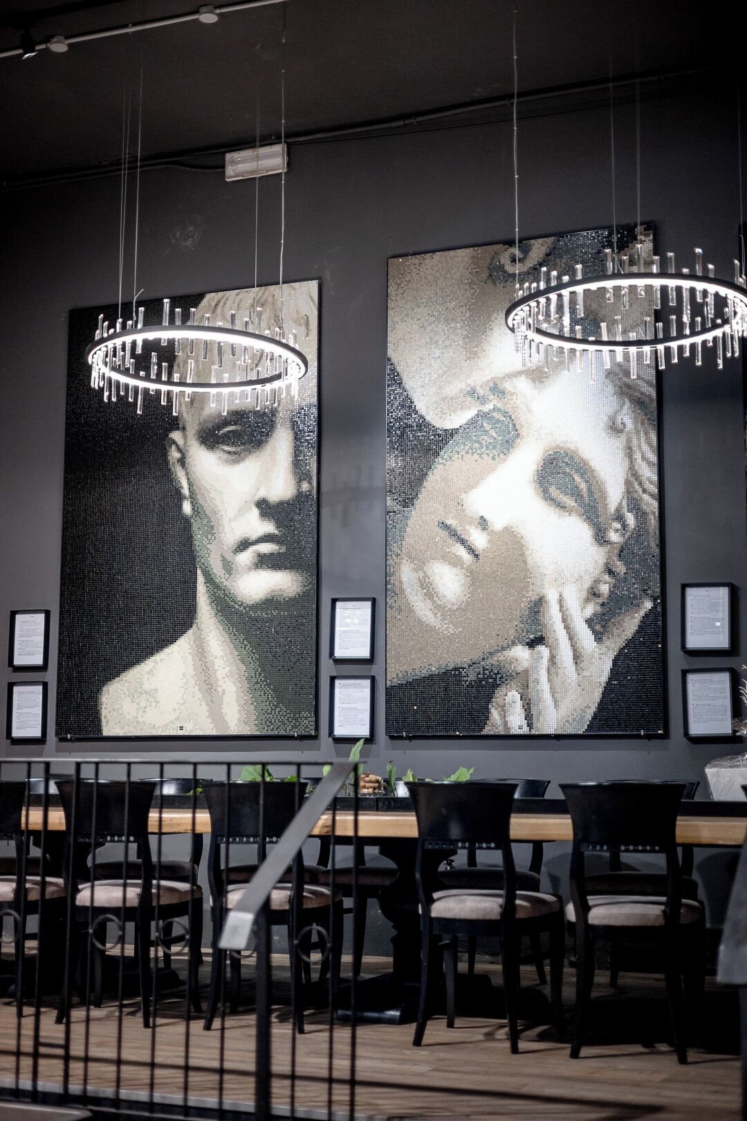 Two large mosaic artworks on a wall, a chandelier above each, and a seating area with tables and chairs in the foreground.