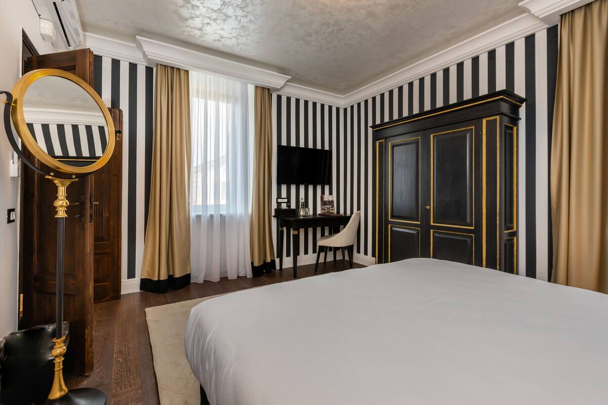 A hotel room with a large bed, a desk with a chair, a wardrobe, a mirror, and a television.