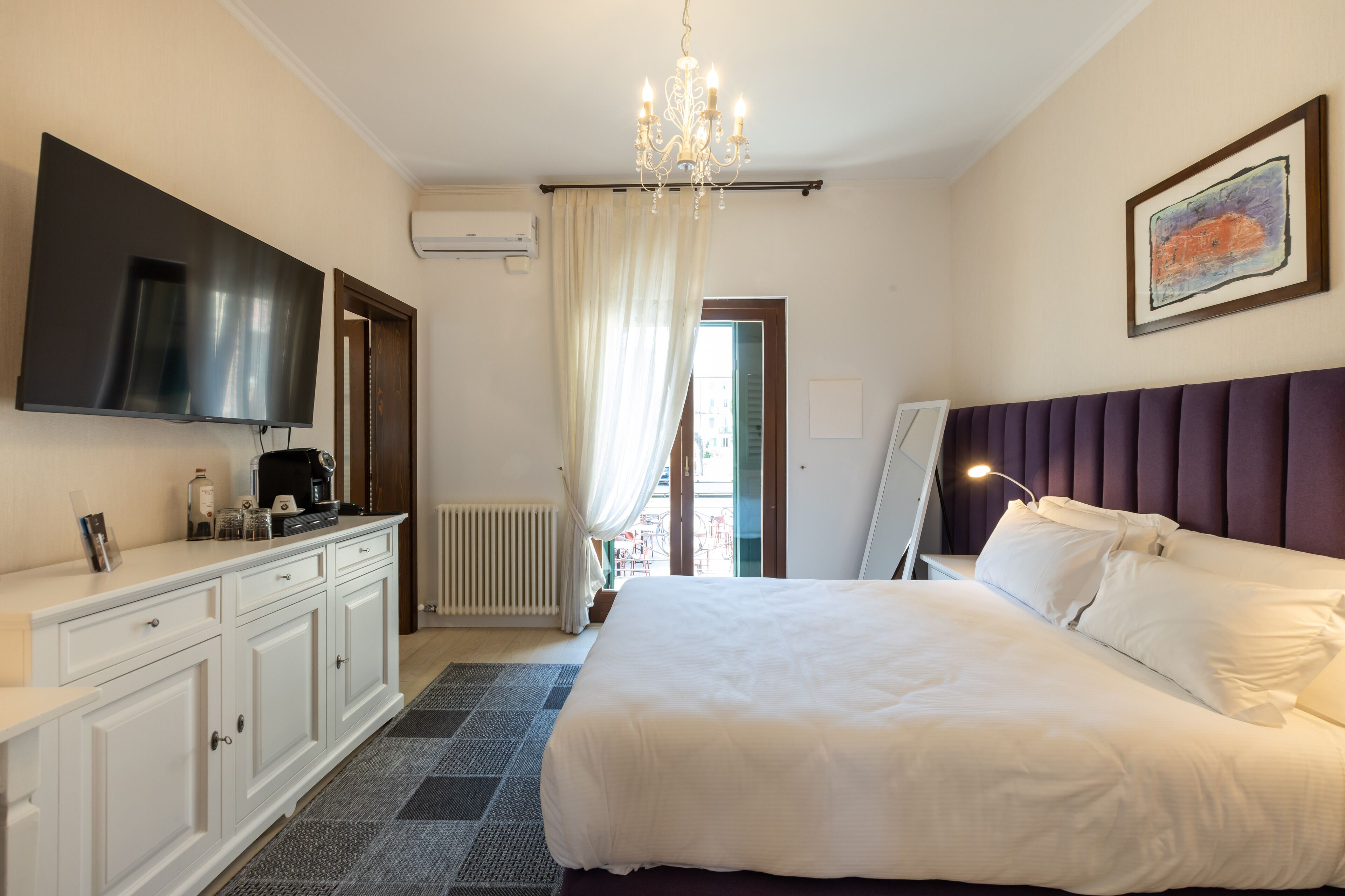Camera d'albergo con un letto grande, una TV a schermo piatto, un lampadario, un quadro e vista sull'esterno.