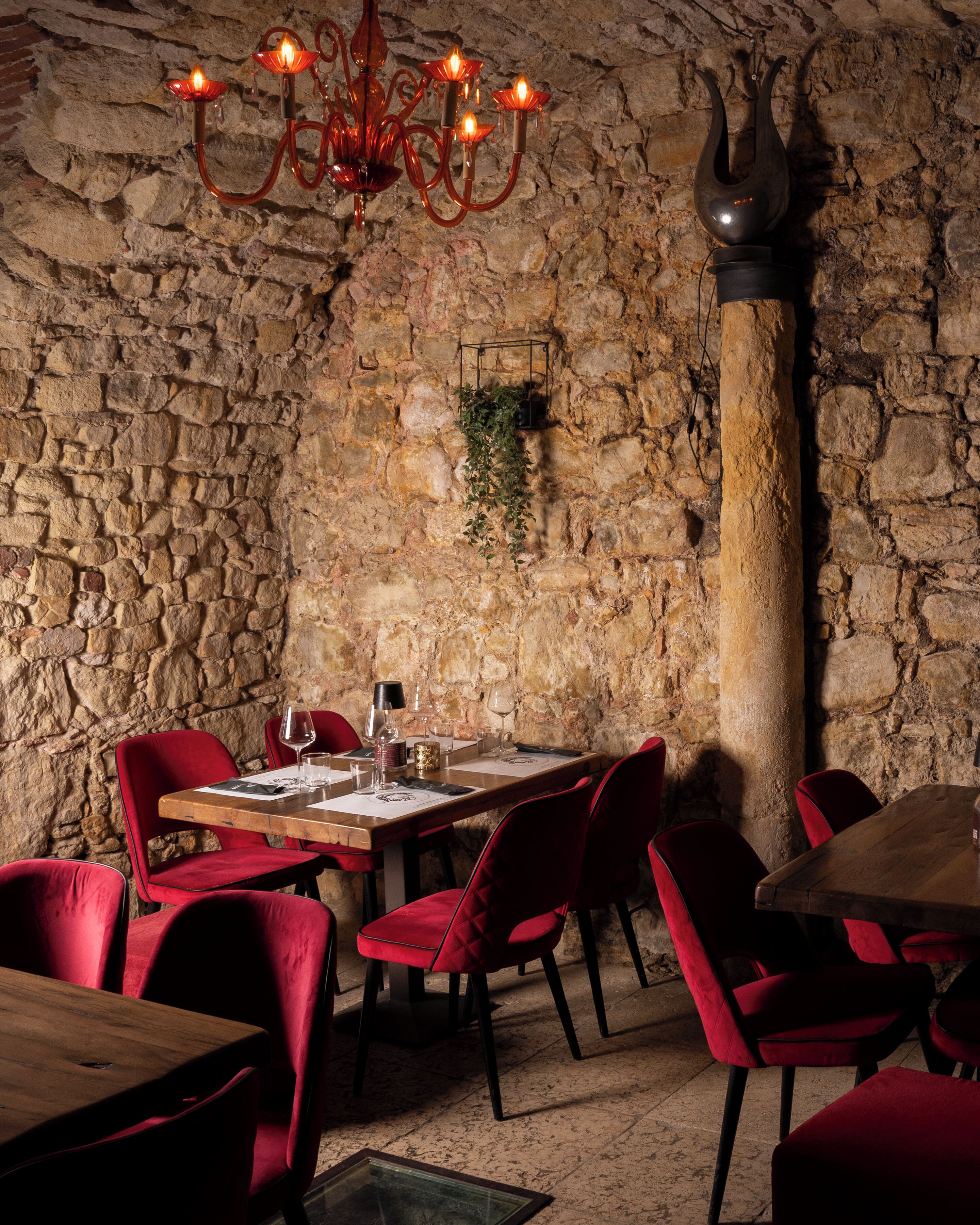 A rustic dining area with stone walls, a wooden table, red upholstered chairs, and a chandelier.