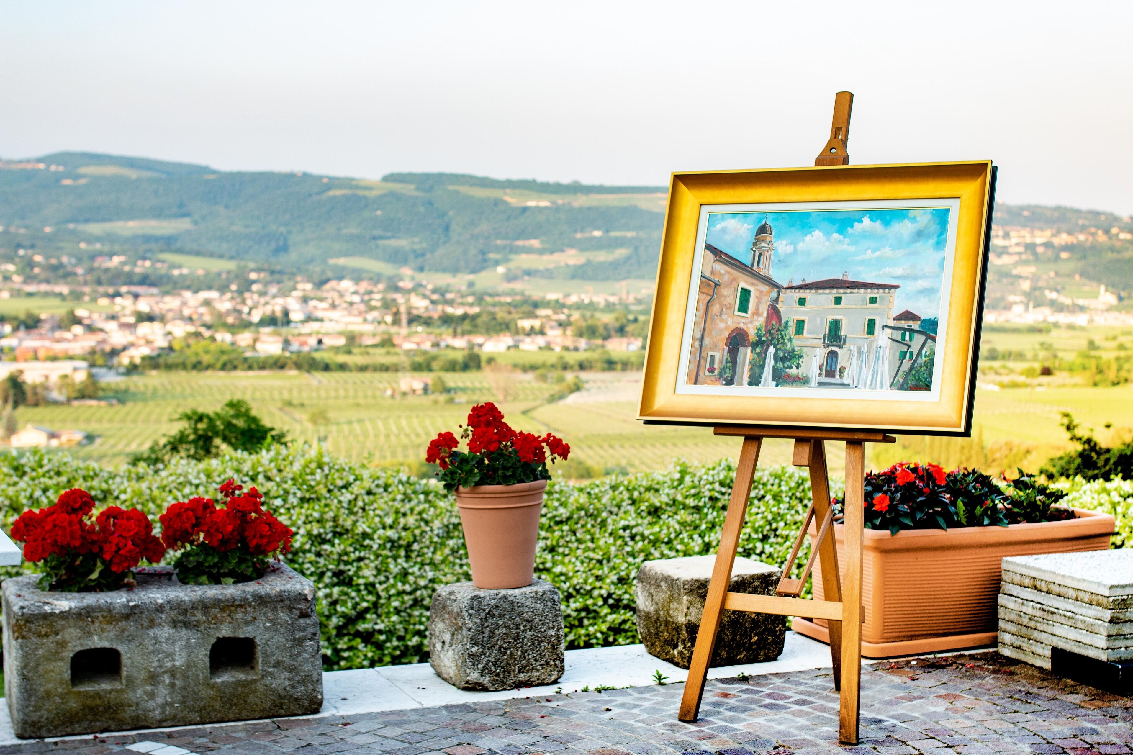 An easel with a painting of a building, surrounded by potted plants and a scenic landscape view.