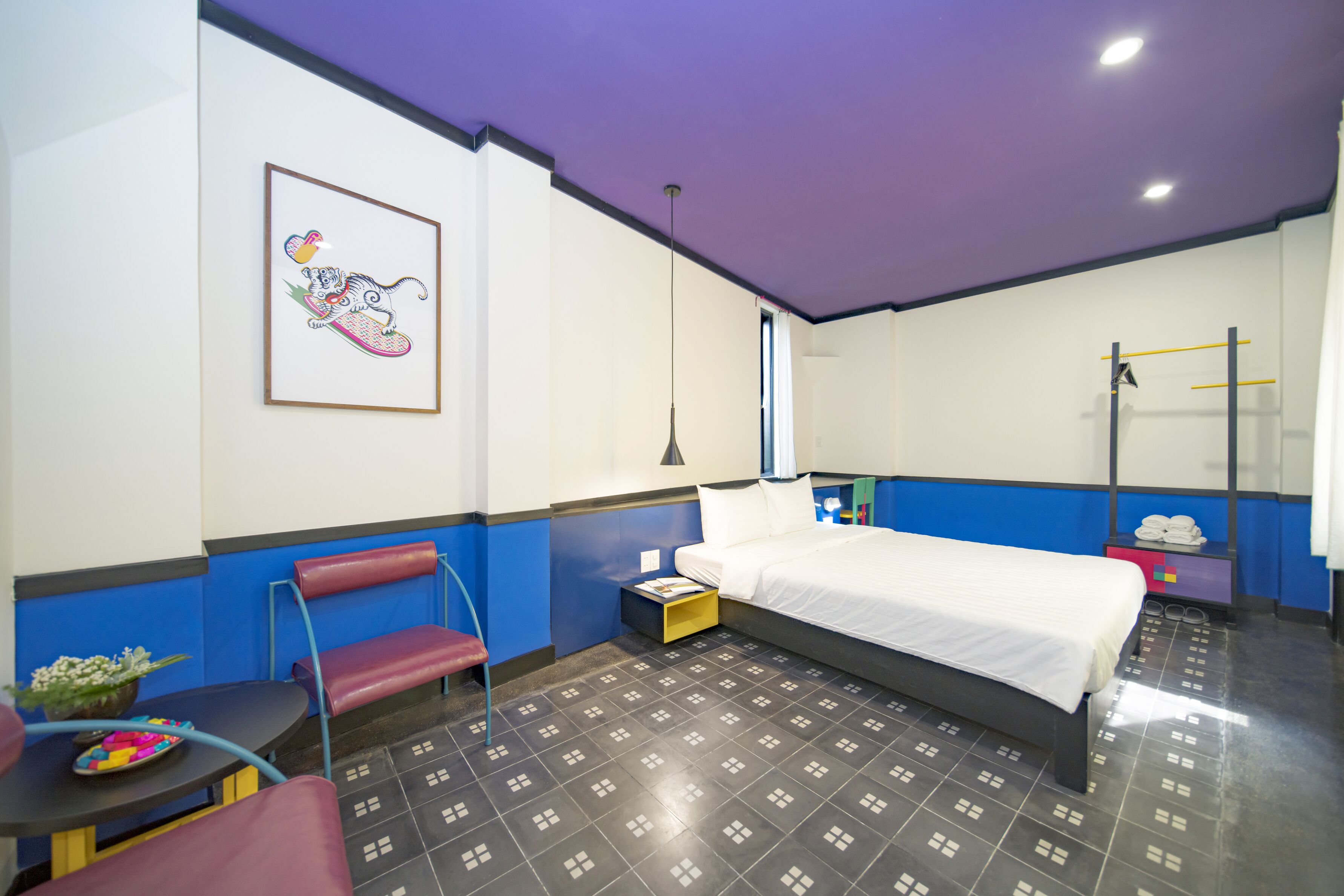 Une chambre d’hôtel avec un lit, une chaise, une petite table sur laquelle est posé un vase de fleurs et un tableau accroché au mur.