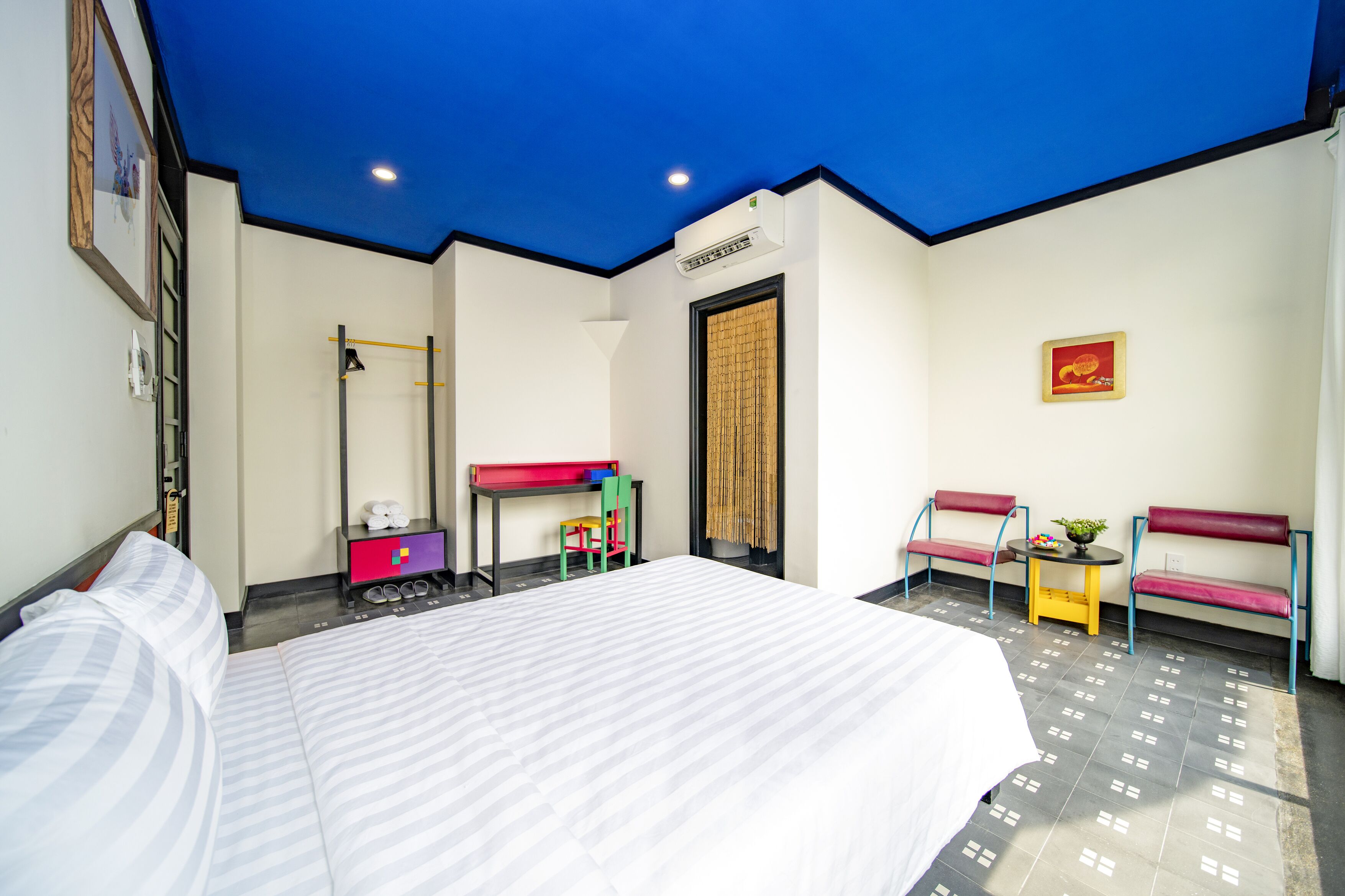 A hotel room with a bed, a desk, two chairs, a small table with a plant, and a ceiling light.