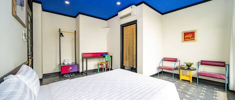 A hotel room with a bed, a desk, two chairs, a small table with a plant, and a ceiling light.