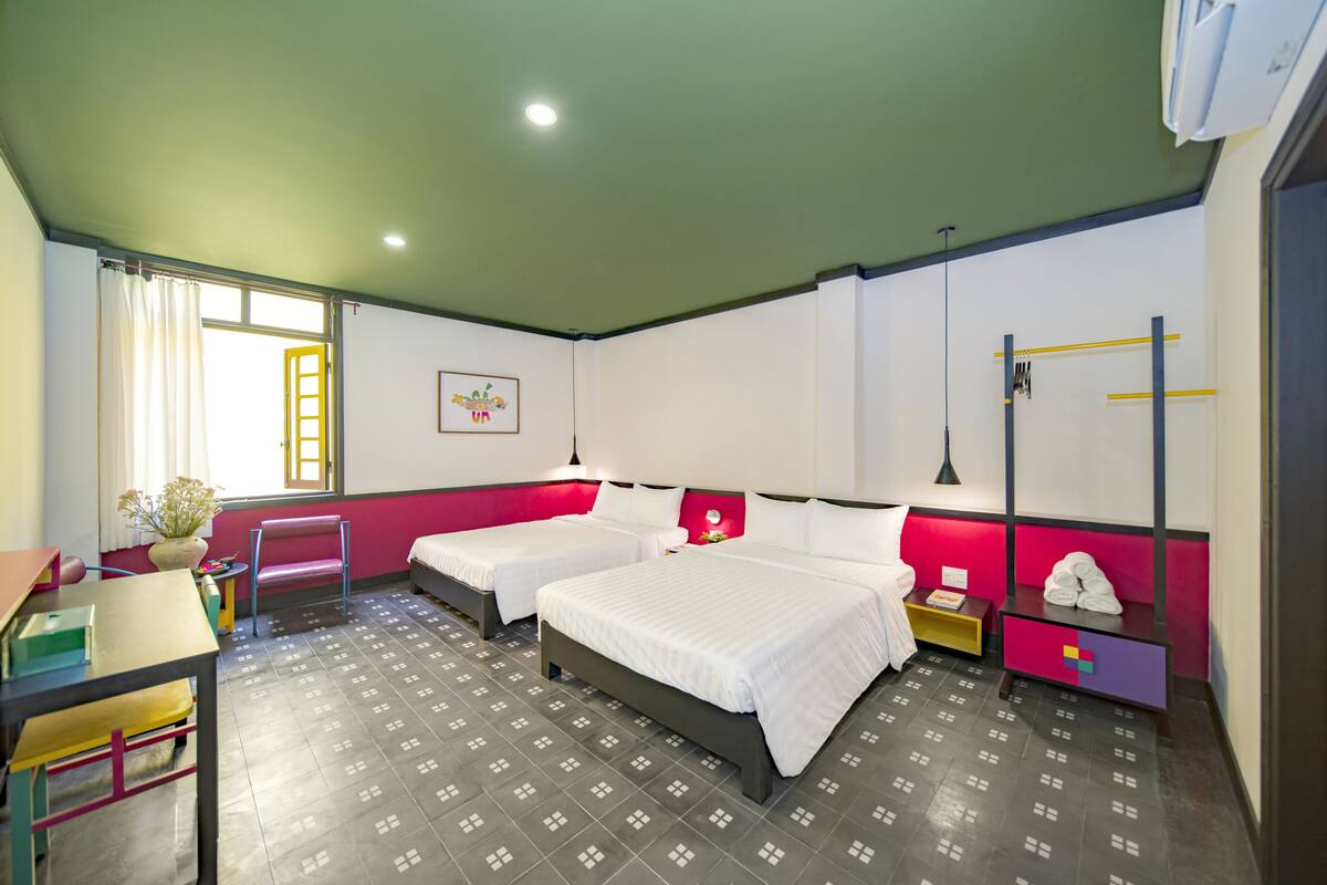 Habitación de hotel con dos camas, un escritorio, una silla, una mesita, una planta en maceta y una ventana con cortina.