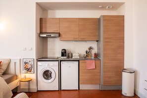 A compact kitchen with a washing machine, a small refrigerator, wooden cabinets, and a trash bin.