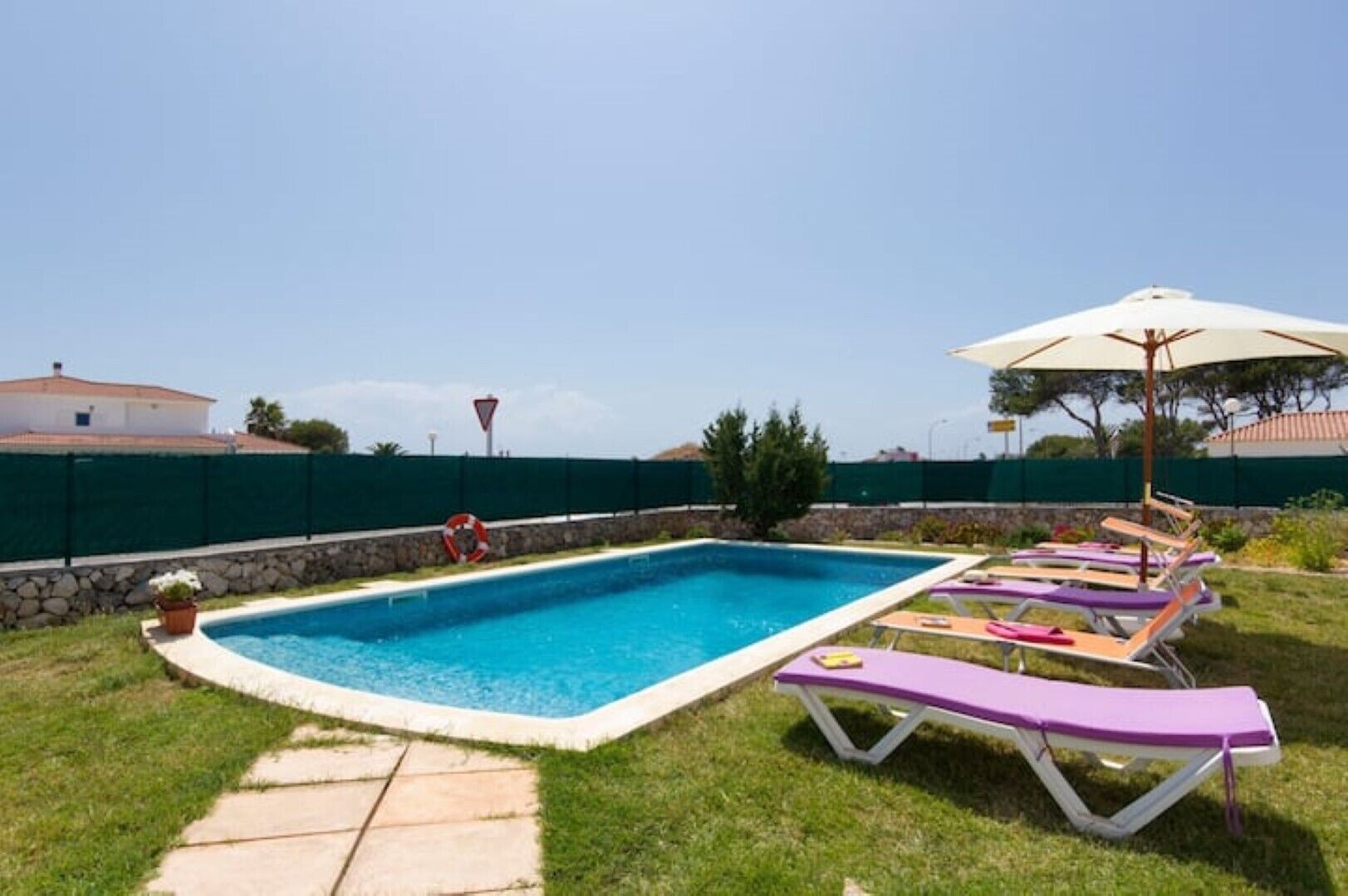 A rectangular swimming pool with a stone border, surrounded by a green fence. There are lounge chairs with purple cushions and a white umbrella.