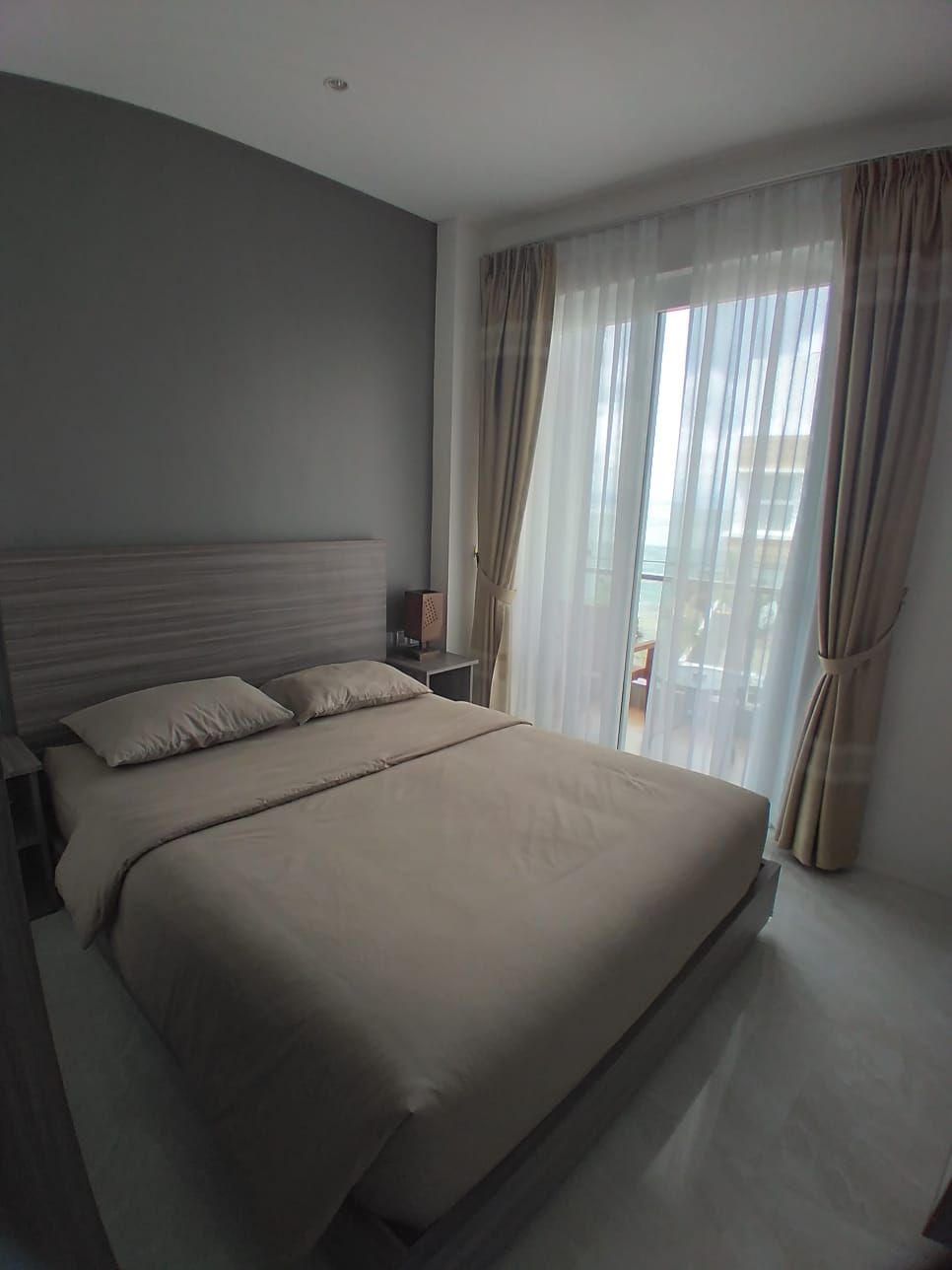A hotel room with a large bed, a nightstand, a lamp, and a view of the outdoors through sheer curtains.