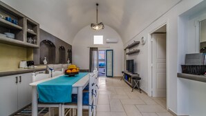 A narrow kitchen with a dining table, chairs, and a fruit bowl. There is a TV and a shelf with items on the right side. - Casa Maritina Private Villa With Jacuzzi (Santorini)