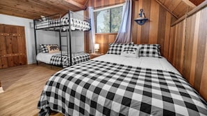 Um quarto estilo cabana com beliche, uma cama grande com roupa de cama xadrez, paredes de madeira, uma janela com cortinas e um ventilador de teto.