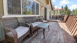 Uma área de estar coberta ao ar livre, com móveis de vime e uma mesa, com vista para um deck com churrasqueira e guarda-sol.