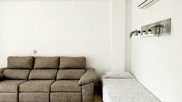 A living room with a sofa and a bed, a wall-mounted air conditioner, and a shelf with plants.