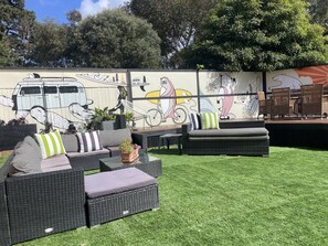 Relax in a serene outdoor seating area with wicker sofas and a chaise lounge, surrounded by lush greenery and a vibrant mural.