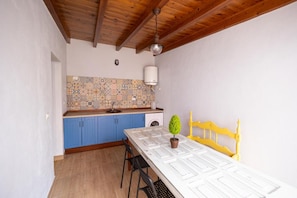 Une cuisine avec des armoires bleues, un pare-feu décoré, un ventilateur de plafond blanc et une table à manger agrémentée d’une plante en pot.