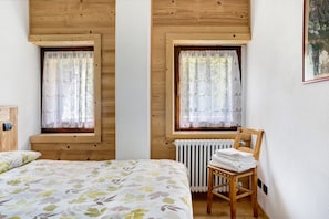 Un dormitorio con cama, dos ventanas con cortinas, un radiador y una silla con toallas.