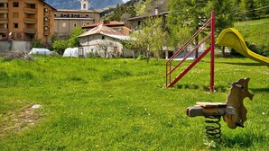 Un campo cubierto de césped con un parque infantil, un edificio con un campanario y montañas al fondo.
