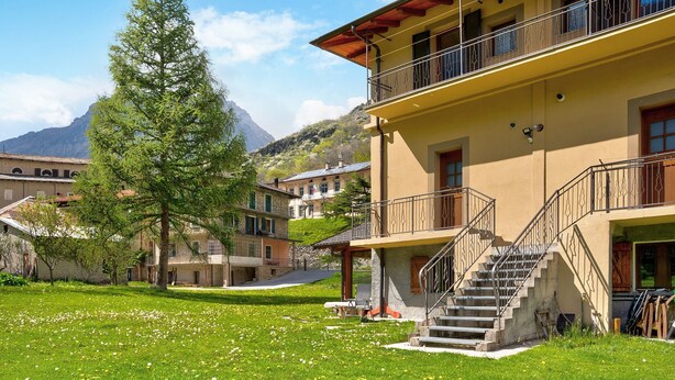 Un bâtiment doté d’un escalier menant à un balcon, entouré d’une zone verdoyante avec des arbres et une montagne en arrière-plan.