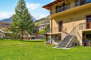 Un bâtiment doté d’un escalier menant à un balcon, entouré d’une zone verdoyante avec des arbres et une montagne en arrière-plan.