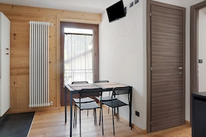Una habitación con suelo de madera, un radiador, una mesa con sillas, un televisor montado en la pared y una puerta.