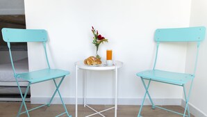Zwei türkisfarbene Stühle mit einem weißen Tisch dazwischen, ein Glas Orangensaft, eine weiße Tasse und eine kleine Vase mit roten Blumen.