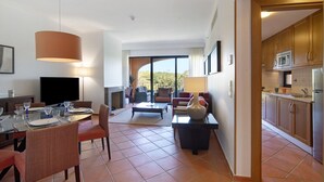 A modern living room with a dining area, a kitchen, and a large window offering a view. - Apartment "T2 Vilasol" with Shared Pool & Wi-Fi (Vilamoura)