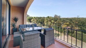 A balcony with wicker furniture, a glass table, and a view of a green landscape. - Apartment "T2 Vilasol" with Shared Pool & Wi-Fi (Vilamoura)