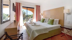 A hotel room with a bed, a desk, a chair, a balcony with curtains, and a view of greenery. - Apartment "T2 Vilasol" with Shared Pool & Wi-Fi (Vilamoura)