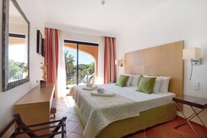 Een hotelkamer met een bed, een bureau, een stoel, een balkon met gordijnen en uitzicht op groen.