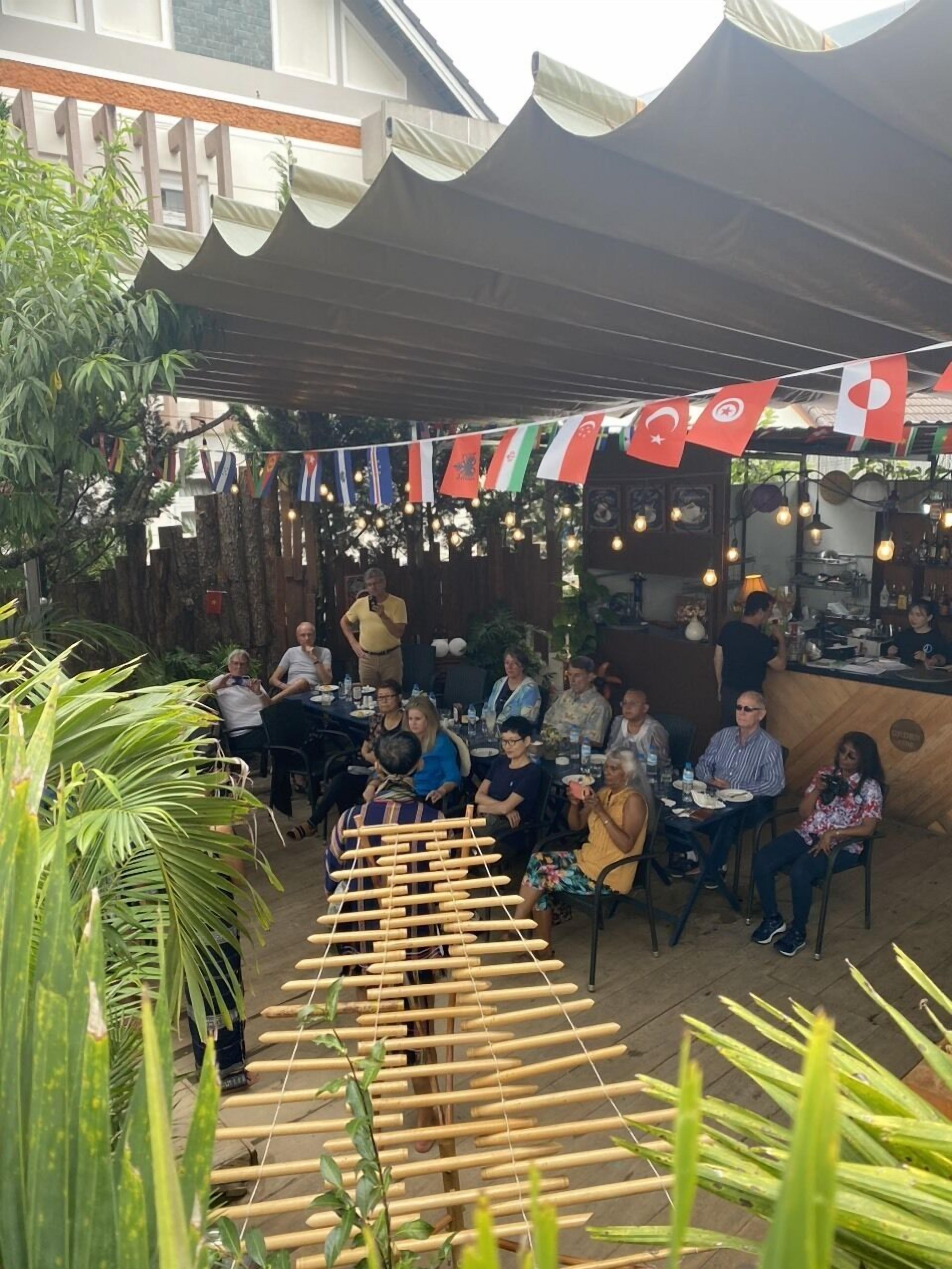 An outdoor dining area with tables and chairs, surrounded by plants and a wooden fence. Flags are strung above, and a bamboo structure is in the foreground.