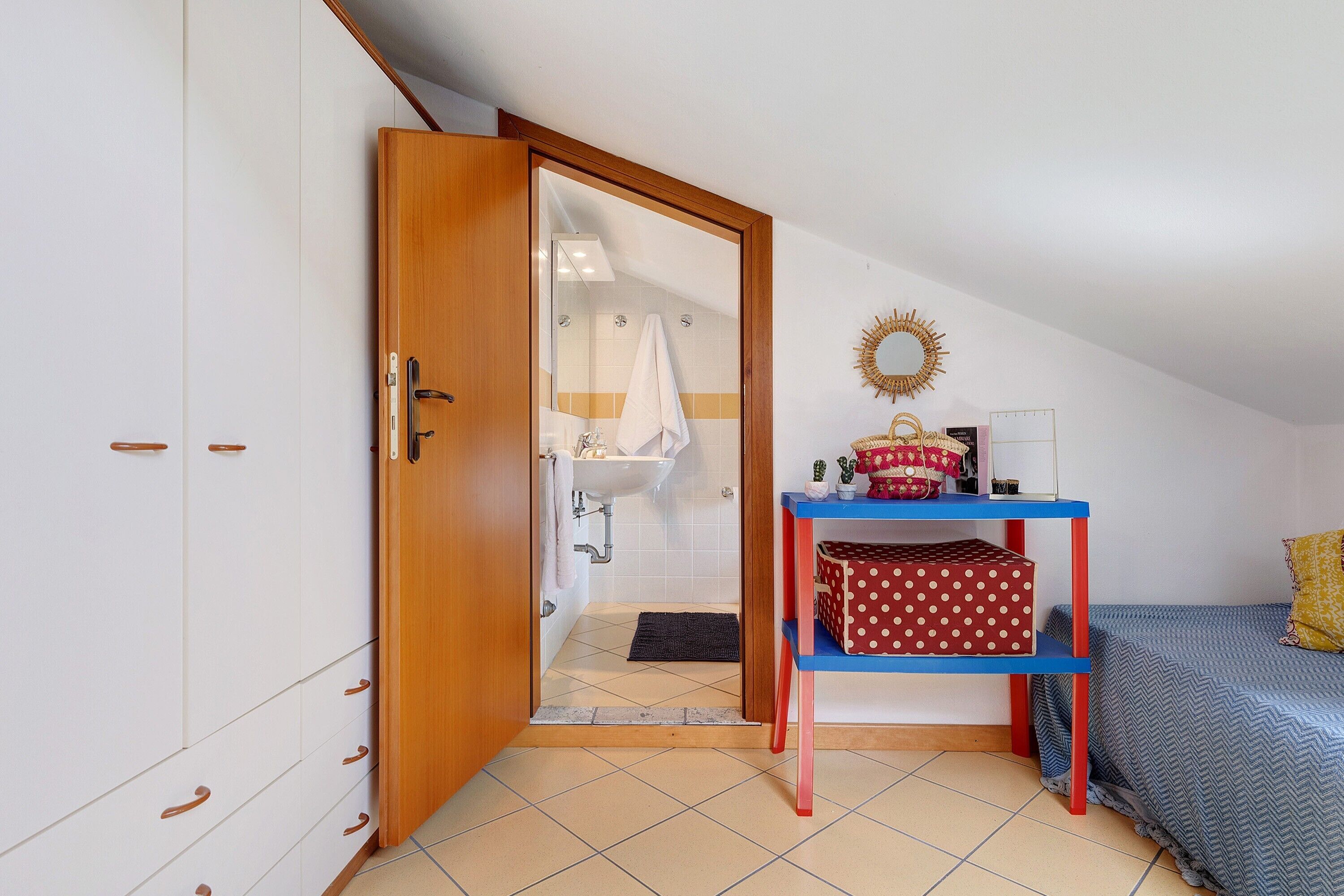 Ein Schlafzimmer mit Bett, Kleiderschrank, Schreibtisch mit Spiegel und einem durchgegangenen Tür angedeuteten Badezimmer.