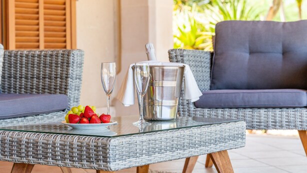 A wicker table with a fruit plate, two wine glasses, and a silver pitcher on a glass table.