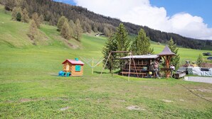 Ein rustikales Holzhaus mit rotem Dach, ein kleines Spielhaus und eine Schaukel in einem grünen Feld mit einem bewaldeten Hügel im Hintergrund.