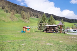 A rustic wooden cabin with a red roof, a small playhouse, and a swing set in a grassy field with a forested hill in the background.